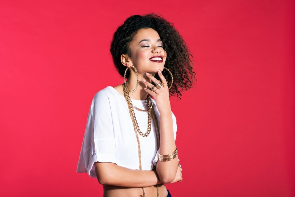 Portrait of happy hip-hop style young latin woman wearing gold jewellery, standing against red background.